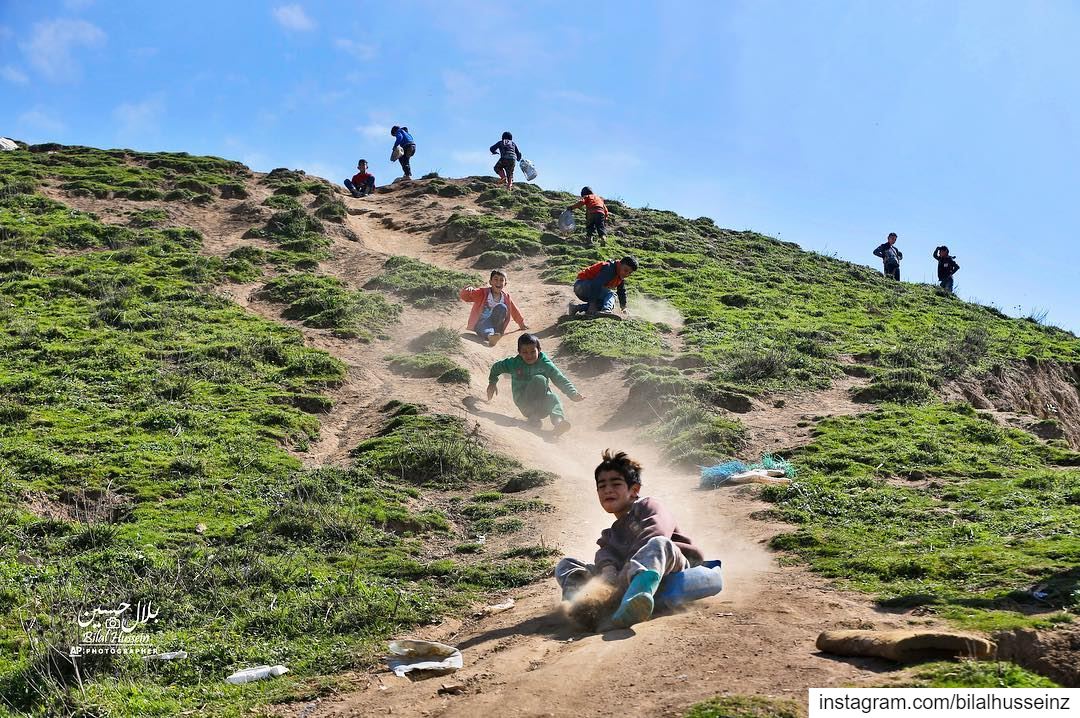 Displaced Syrian children slide down a hill, above a refugee camp in the...