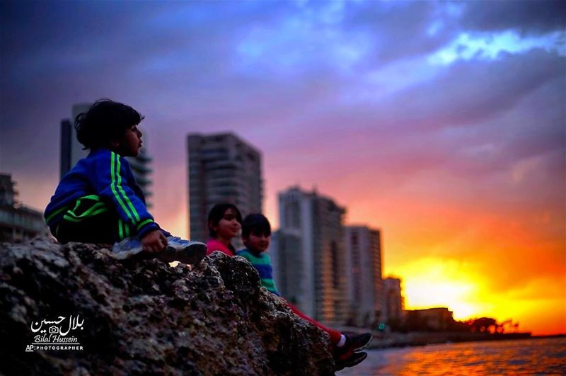 Children sit on a rocky area along the coastline as the sunset over the...