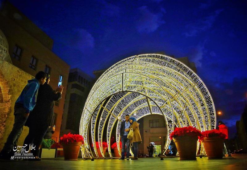 Two people take a photo of a couple near Christmas decorations in downtown...